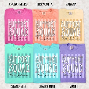 May include: Six t-shirts in crunchberry, terracotta, banana, island reef, chalky mint, and violet colors. Each shirt features a vertical striped background, the text 'SUPPORT SQUAD' in white, and the personalized name 'Mrs. Johnson' in black script.