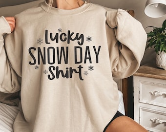 May include: A beige sweatshirt with the text "Lucky Snow Day Shirt" printed on the front. The text is in black and surrounded by small snowflake graphics.