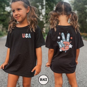 May include: Black t-shirt with a peace sign graphic on the back and the word "USA" on the front. The peace sign is red, white, and blue with stars and stripes.