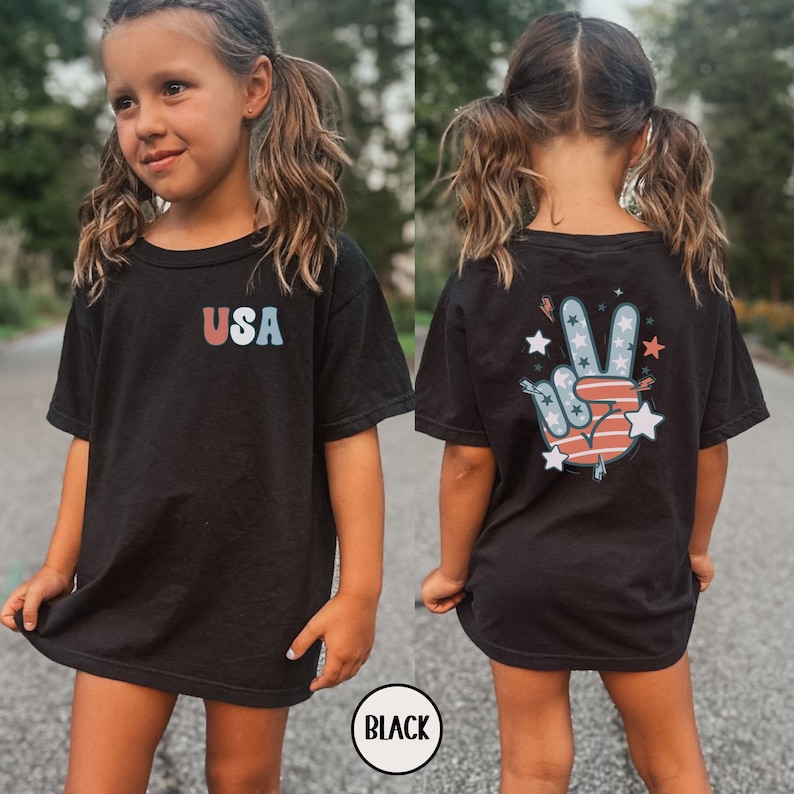 May include: Black t-shirt with a peace sign graphic on the back and the word "USA" on the front. The peace sign is red, white, and blue with stars and stripes.