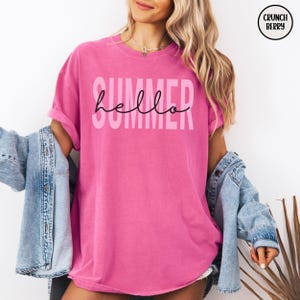 May include: A bright pink t-shirt with the word "SUMMER" in light pink block letters and the word "hello" in black cursive script. The shirt is part of a summer clothing collection.