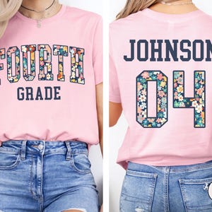 May include: A light pink t-shirt with the words "FOURTH GRADE" in a floral design. The back of the shirt has the name "JOHNSON" and the number "04" in a floral design. The shirt is paired with blue denim jeans.