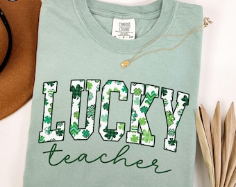 Puede incluir: Una camiseta verde claro con las palabras "LUCKY teacher" impresas en blanco con un patrón de trébol.