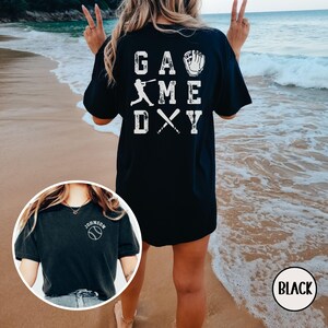 May include: Black t-shirt with white text that says "Game Day" with a baseball player, bat and glove graphic. The shirt is oversized and worn by a person standing on a beach.