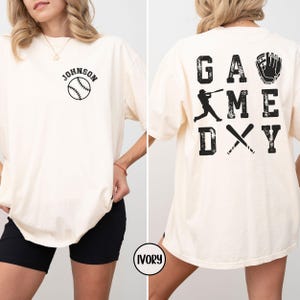 May include: Ivory t-shirt with a baseball graphic and the text "JOHNSON" on the front. The back of the shirt has the text "GAME DAY" with a baseball player graphic and a baseball glove graphic.