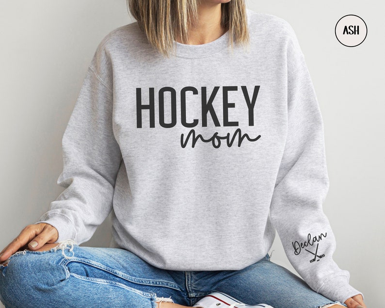 May include: A grey sweatshirt with the text "HOCKEY mom" in black. The sweatshirt has a small logo on the sleeve with the text "Declan" and a hockey stick graphic.