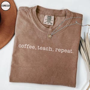 May include: A brown Comfort Colors t-shirt with the text "coffee, teach, repeat" in white. A silver necklace with a small pendant is draped across the shirt. The shirt is a small size.