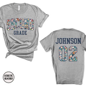 Puede incluir: Camiseta gris jaspeado con "SECOND GRADE" en un dise&ntilde;o floral en la parte delantera. La parte trasera de la camiseta muestra "JOHNSON" y el n&uacute;mero "02" en un patr&oacute;n floral a juego. La camiseta est&aacute; hecha de material Athletic Heather.
