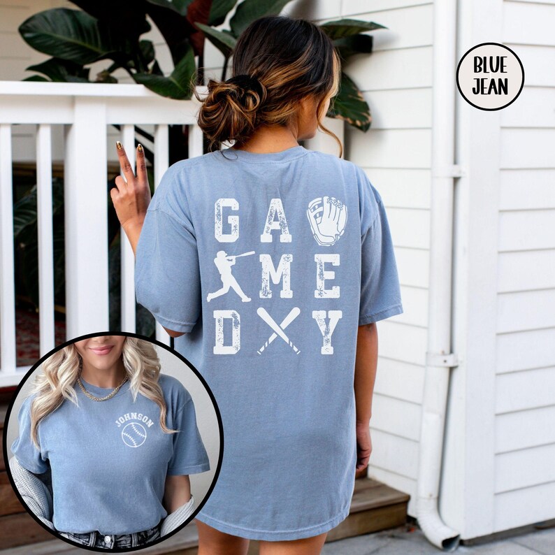 May include: A blue jean t-shirt with the words "GAME DAY" in white letters on the back. The letters are arranged in a grid pattern with a baseball player, a baseball bat, and a baseball glove in the spaces between the letters.
