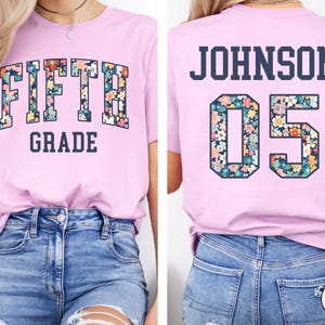 Op de afbeelding: Een lichtpaars T-shirt met de tekst "FIFTH GRADE" in bloemenletters op de voorkant. De achterkant van het shirt toont "JOHNSON 05" in een vergelijkbaar bloemenontwerp. Het shirt is gemaakt van zacht materiaal en geschikt voor dagelijks gebruik.