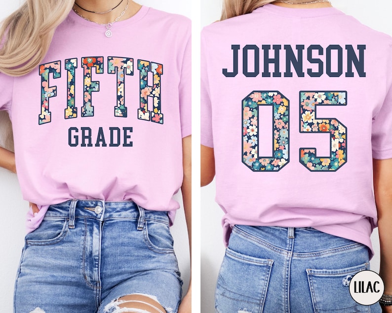 Op de afbeelding: Een lichtpaars T-shirt met de tekst "FIFTH GRADE" in bloemenletters op de voorkant. De achterkant van het shirt toont "JOHNSON 05" in een vergelijkbaar bloemenontwerp. Het shirt is gemaakt van zacht materiaal en geschikt voor dagelijks gebruik.