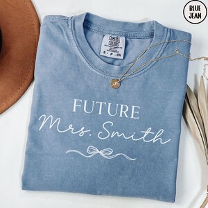 May include: A light blue t-shirt with the text "FUTURE Mrs. Smith" in white lettering. The shirt has a distressed look and a small bow underneath the text.