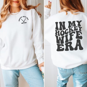 Pu&ograve; includere: Felpa bianca con il testo "In My Hockey Wife Era" in un font retr&ograve; sul retro e una piccola grafica di bastone da hockey sul davanti.
