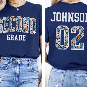 Puede incluir: Camiseta azul marino con "SECOND GRADE" en letras florales en la parte delantera. La parte trasera muestra "JOHNSON 02" con n&uacute;meros estampados florales. Una prenda casual con tem&aacute;tica escolar.