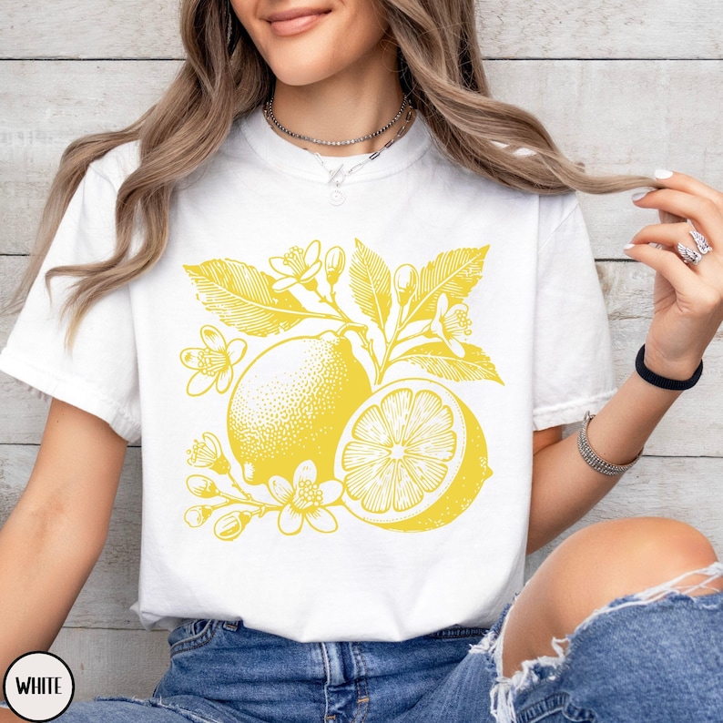 Op de afbeelding: Wit T-shirt met een gele citroenprint. De citroen is omringd door bladeren en bloemen.
