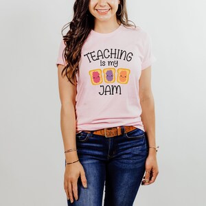 Peut inclure: Un t-shirt rose avec le texte "TEACHING is my JAM" et un graphique de trois tranches de pain grill&eacute; avec de la gel&eacute;e de diff&eacute;rentes couleurs.