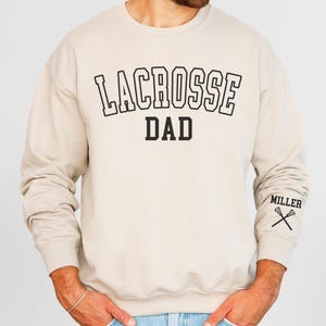 May include: A sand-colored sweatshirt with the text "LACROSSE DAD" in black. The left sleeve has the text "MILLER" and two lacrosse sticks crossed.