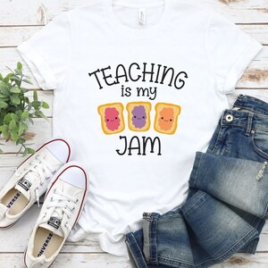 Peut inclure: T-shirt blanc avec le texte "TEACHING is my JAM" et trois images de toasts de dessin anim&eacute; avec de la gel&eacute;e de diff&eacute;rentes couleurs. Le toast a un visage heureux.