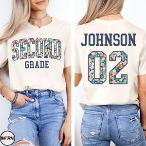 May include: Cream-colored t-shirt with "SECOND GRADE" in floral letters on the front. The back features "JOHNSON" and the number "02" in a floral pattern. The shirt is paired with blue jeans.