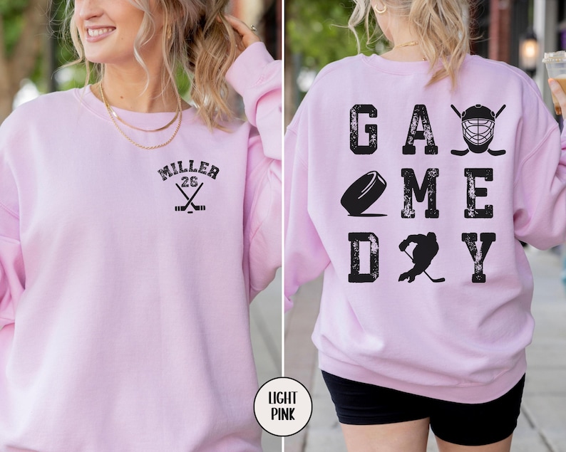 May include: Light pink sweatshirt with the text "GAME DAY" on the back and a hockey stick and puck graphic. The front of the sweatshirt has the text "MILLER 26" with a hockey stick graphic.
