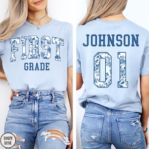 以下が含まれることがあります： 前面に花柄のデザインで「FIRST GRADE」と書かれた水色のTシャツ。背中にはお揃いの花柄で「JOHNSON 01」と表示されています。Tシャツはベビーブルー色です。