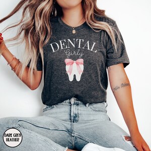 May include: Dark grey heather t-shirt with the text "Dental Girly" in white. A pink bow is on a white tooth graphic.