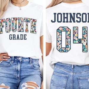 May include: White t-shirt with "FOURTH GRADE" in floral letters on the front. The back of the shirt displays "JOHNSON" and the number "04" in a floral pattern. The shirt is paired with blue jeans.