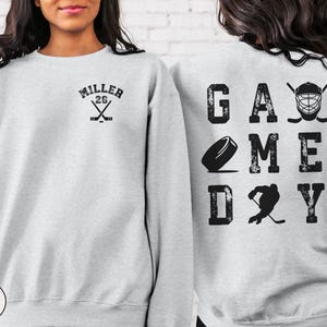 May include: A gray sweatshirt with a hockey-themed design. The front of the sweatshirt has a hockey stick and the text "MILLER 26". The back of the sweatshirt has the text "GAME DAY" with a hockey puck, a hockey player, and a hockey helmet.