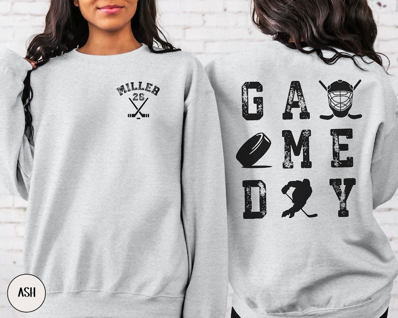 May include: A gray sweatshirt with a hockey-themed design. The front of the sweatshirt has a hockey stick and the text "MILLER 26". The back of the sweatshirt has the text "GAME DAY" with a hockey puck, a hockey player, and a hockey helmet.