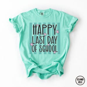 May include: A light turquoise t-shirt with the text "HAPPY LAST DAY OF SCHOOL" in black, with pink star accents. The text is set against a background of vertical stripes. The shirt has short sleeves and a crew neck.
