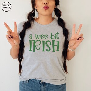 以下が含まれることがあります： 緑色の「a wee bit IRISH」の文字が入ったグレーのTシャツ。クルーネックと半袖です。ブランドロゴ「ATHLETIC HEATHER」が左上に円で表示されています。モデルはピースサインをしています。