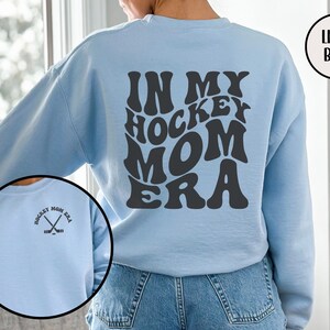 May include: Light blue sweatshirt with the text "In My Hockey Mom Era" in a retro font on the back. The front of the sweatshirt has a small graphic of two hockey sticks crossed with the text "Hockey Mom Era".
