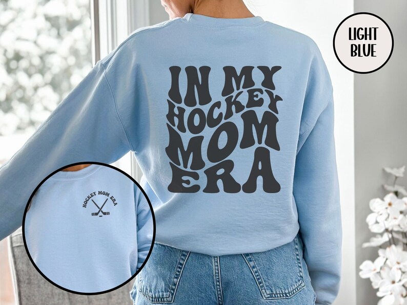 May include: Light blue sweatshirt with the text "In My Hockey Mom Era" in a retro font on the back. The front of the sweatshirt has a small graphic of two hockey sticks crossed with the text "Hockey Mom Era".