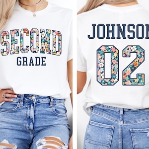 Puede incluir: Camiseta blanca con "SECOND GRADE" en estampado floral en la parte delantera. La parte trasera de la camiseta muestra "JOHNSON" y el n&uacute;mero "02", tambi&eacute;n con un patr&oacute;n floral. Estilo casual.