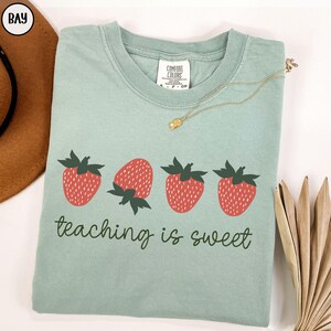 May include: A light green t-shirt with a crew neck, featuring four red strawberries with green leaves. The phrase "teaching is sweet" is printed below the strawberries in a cursive font. A gold necklace is draped across the shirt.