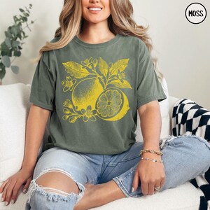 Op de afbeelding: Een vrouw draagt een groen T-shirt met een gele citroenprint. Het shirt heeft een distressed look en is oversized. De vrouw draagt een blauwe jeans en heeft haar haar naar achteren getrokken.
