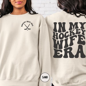 Pu&ograve; includere: Una felpa color crema con la scritta "Hockey Wife Era" in nero sul retro. Il testo &egrave; in un font retr&ograve;. La parte anteriore della felpa ha un piccolo grafico nero di bastoni da hockey incrociati con un disco.
