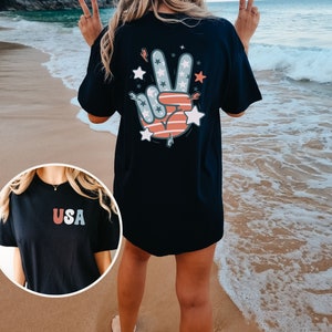 May include: Black t-shirt with a peace sign graphic featuring red, white, and blue stars and stripes. The back of the shirt has the word "USA" in red, white, and blue.