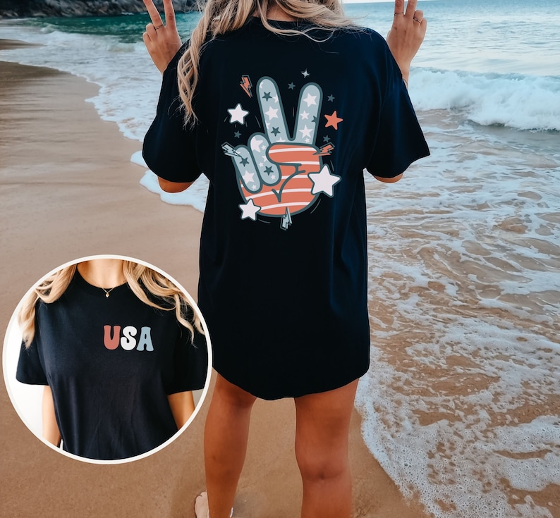 May include: Black t-shirt with a peace sign graphic featuring red, white, and blue stars and stripes. The back of the shirt has the word "USA" in red, white, and blue.