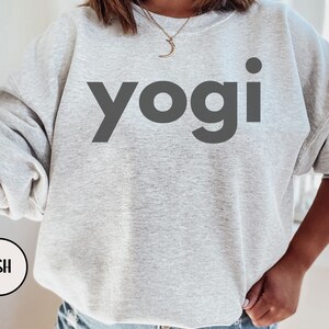 Może przedstawiać: Szara bluza z nadrukiem słowa "yogi" w kolorze czarnym na przodzie. Bluza wykonana jest z miękkiego, wygodnego materiału i ma luźny kr&oacute;j.