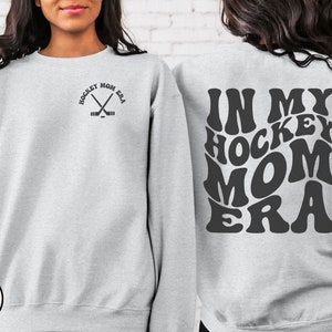 May include: A gray sweatshirt with the text "Hockey Mom Era" in a retro font on the back. The front of the sweatshirt has a small graphic of two hockey sticks crossed with the text "Hockey Mom Era" above it.