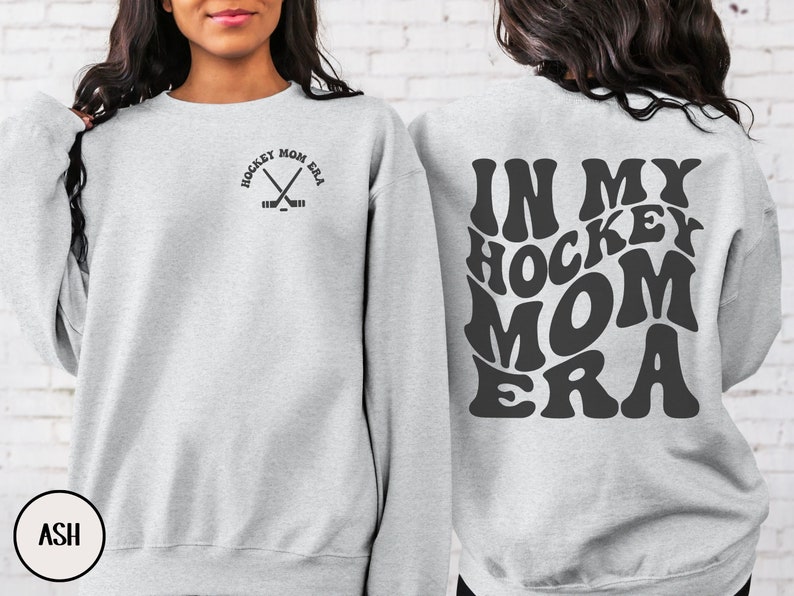 May include: A gray sweatshirt with the text "Hockey Mom Era" in a retro font on the back. The front of the sweatshirt has a small graphic of two hockey sticks crossed with the text "Hockey Mom Era" above it.