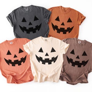 May include: A collection of five short-sleeved t-shirts in various colors, each featuring a black jack-o'-lantern face design. The shirts are in shades of gray, orange, peach, cream, and brown. The design includes triangular eyes and a jagged mouth.