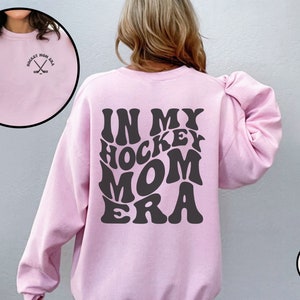 May include: Light pink sweatshirt with the text "In My Hockey Mom Era" in a retro font on the back. The front of the sweatshirt has a small embroidered design of hockey sticks.