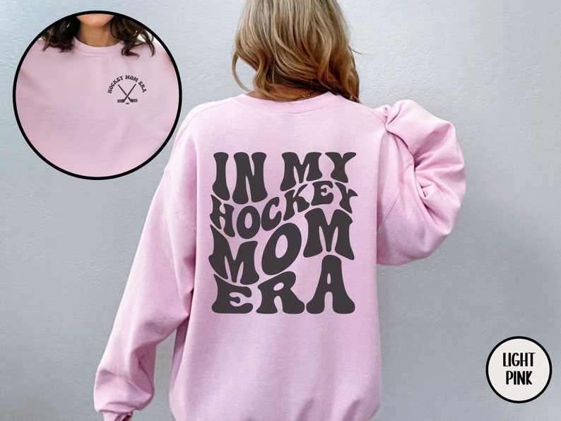 May include: Light pink sweatshirt with the text "In My Hockey Mom Era" in a retro font on the back. The front of the sweatshirt has a small embroidered design of hockey sticks.