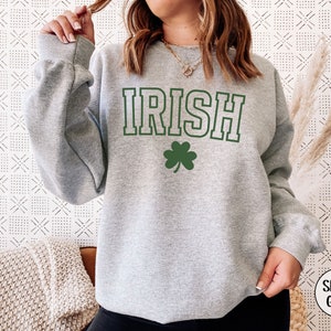 Op de afbeelding: Een grijs sweatshirt met het woord "IRISH" in groene letters en een groene klaver. Het sweatshirt is een ronde hals en heeft een ontspannen pasvorm.