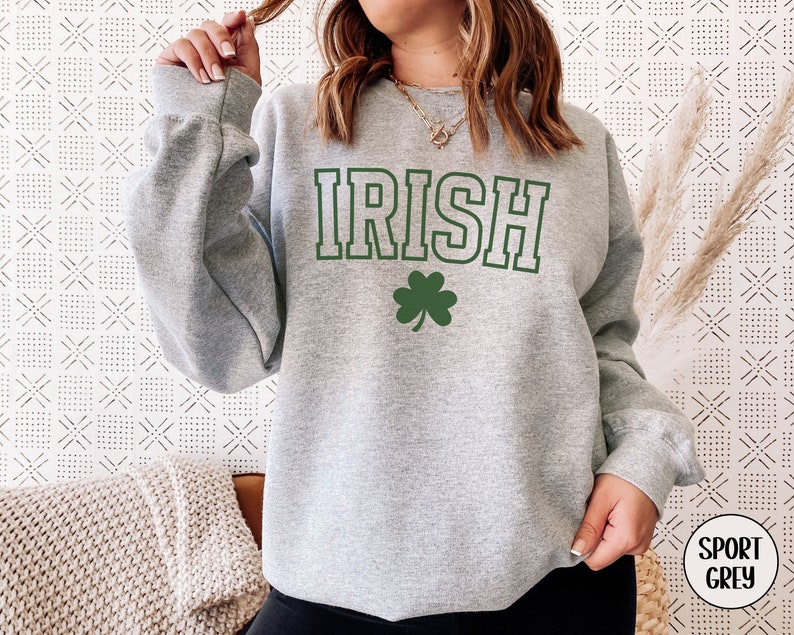 Op de afbeelding: Een grijs sweatshirt met het woord "IRISH" in groene letters en een groene klaver. Het sweatshirt is een ronde hals en heeft een ontspannen pasvorm.