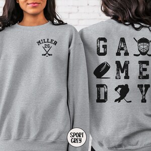 May include: Gray sweatshirt with the text "GAME DAY" in black with hockey-themed graphics. The back of the sweatshirt has the text "MILLER 26" with crossed hockey sticks.