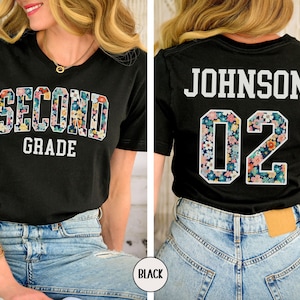 Puede incluir: Camiseta negra con las palabras "SECOND GRADE" en un dise&ntilde;o floral en la parte delantera. La parte trasera de la camiseta muestra el nombre "JOHNSON" y el n&uacute;mero "02" en un patr&oacute;n floral. La camiseta se usa con vaqueros azules.