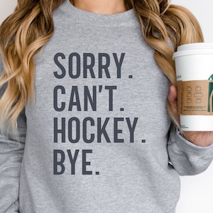 May include: Gray sweatshirt with the text "SORRY. CAN'T. HOCKEY. BYE." printed on the front. A person is holding a white coffee cup with a green logo.